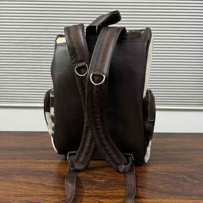 A dark brown textured leather backpack shown from the back, resting on a wooden surface in front of a window with horizontal blinds.