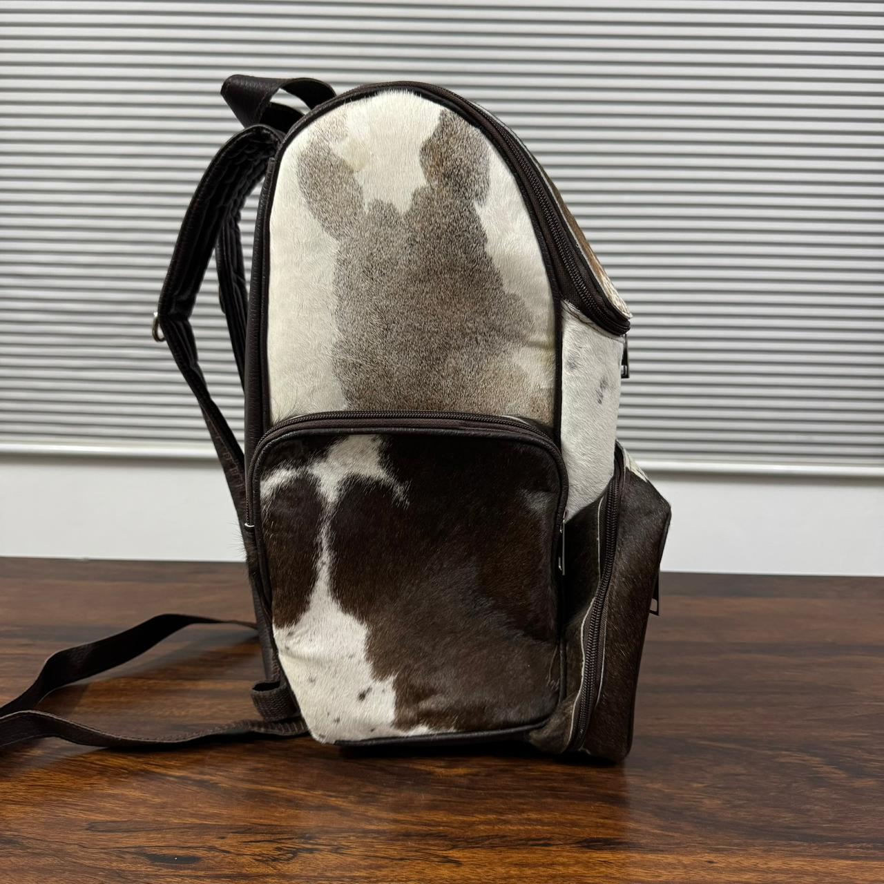 A cowhide backpack with dark brown and white patches, featuring dark brown leather trim, zippers, and straps, resting on a wooden table in front of a gray slatted window.