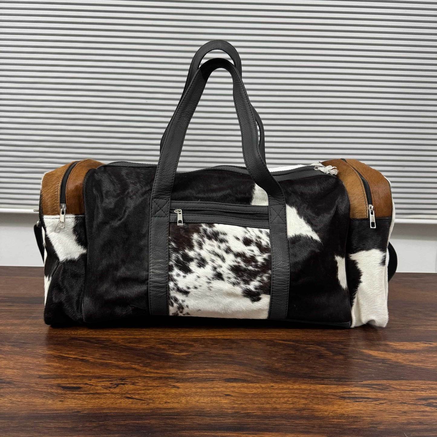 A duffle bag with a black and white cowhide pattern, brown leather side panels, and black faux-leather handles, sitting on a wooden table in front of a gray slatted background.