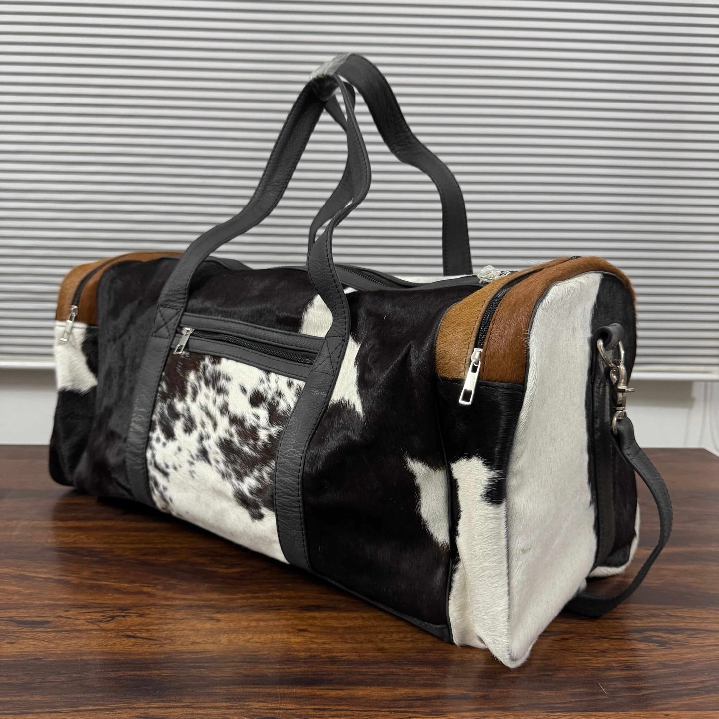 A cowhide duffel bag with black and white spots, brown leather end panels, and black straps, sitting on a wooden table in front of a gray slatted window.