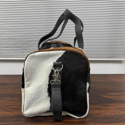A small duffel bag with a black and white cowhide pattern, brown leather trim, and black leather handles, standing on a wooden surface in front of a window with horizontal blinds.