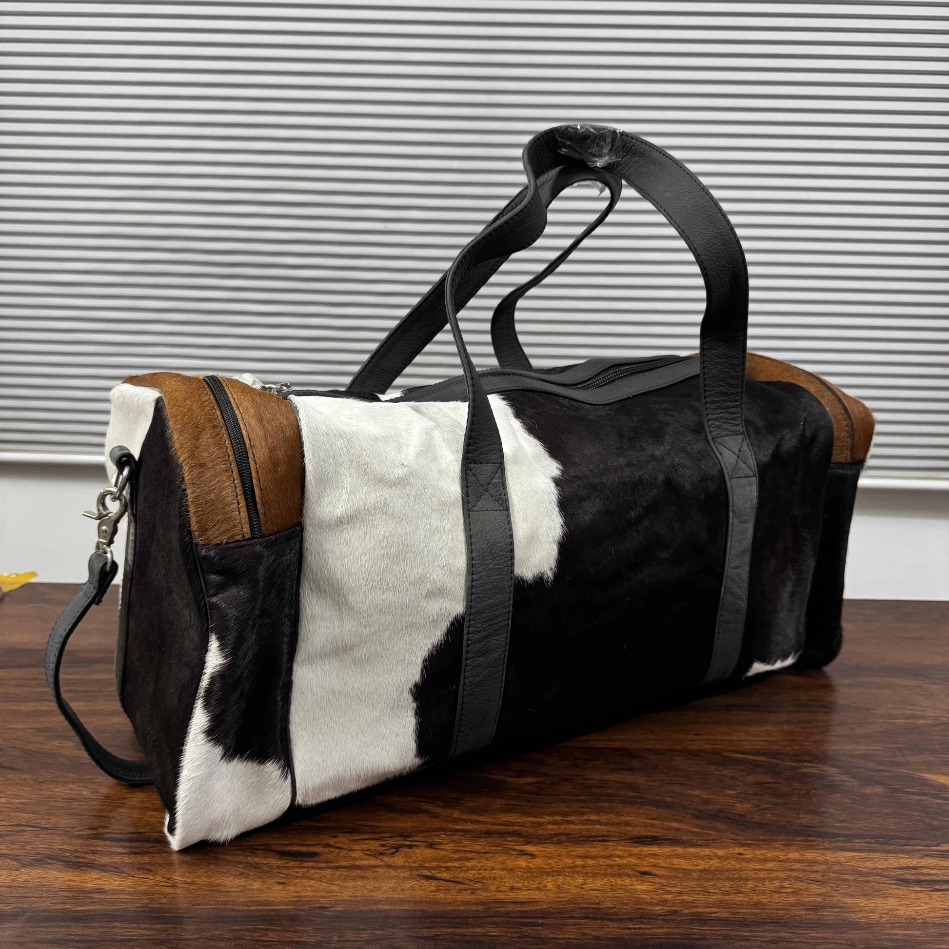 A duffle bag with a black and white cowhide pattern, brown leather trim, and black straps, sitting on a wooden table in front of a gray slatted wall.