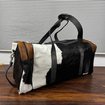 A duffle bag with a black and white cowhide pattern, brown leather trim, and black straps, sitting on a wooden table in front of a gray slatted wall.