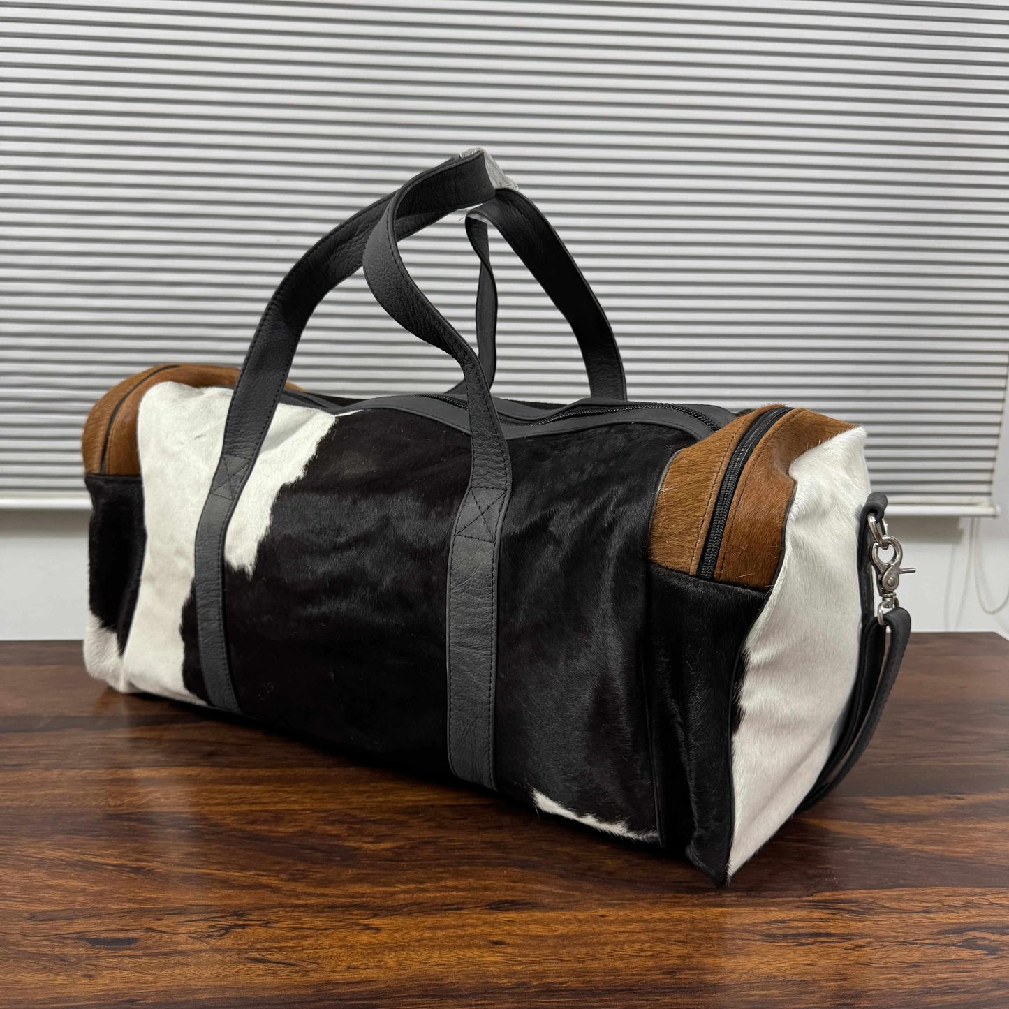 A duffle bag made of black and white cowhide with brown leather trim and black handles, resting on a wooden table in front of a window with horizontal blinds.
