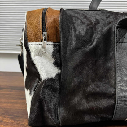 A close-up of a handbag featuring a black and white cowhide pattern, a brown hair-on-hide panel, and dark gray leather trim, resting on a wooden surface.