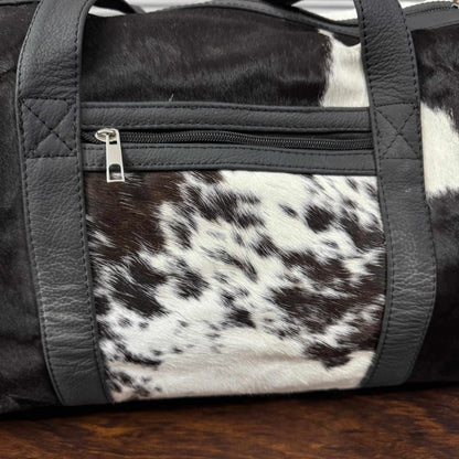 A close-up of a duffel bag featuring a black and white cowhide pattern on the front pocket and handles, contrasted with smooth black faux-leather trim and a silver zipper pull.