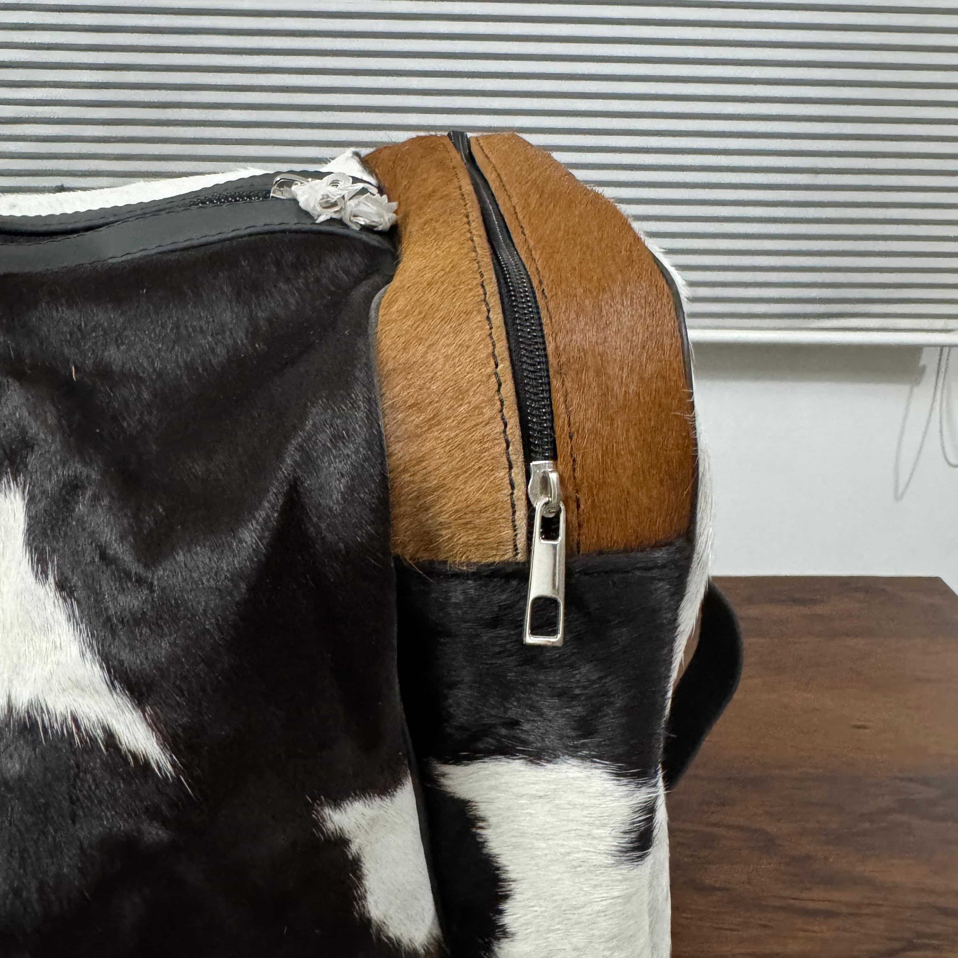 Close-up of a cowhide backpack or duffel bag showing a black-and-white patterned hide, a tan side panel, and a black zipper with a silver pull on a tabletop.