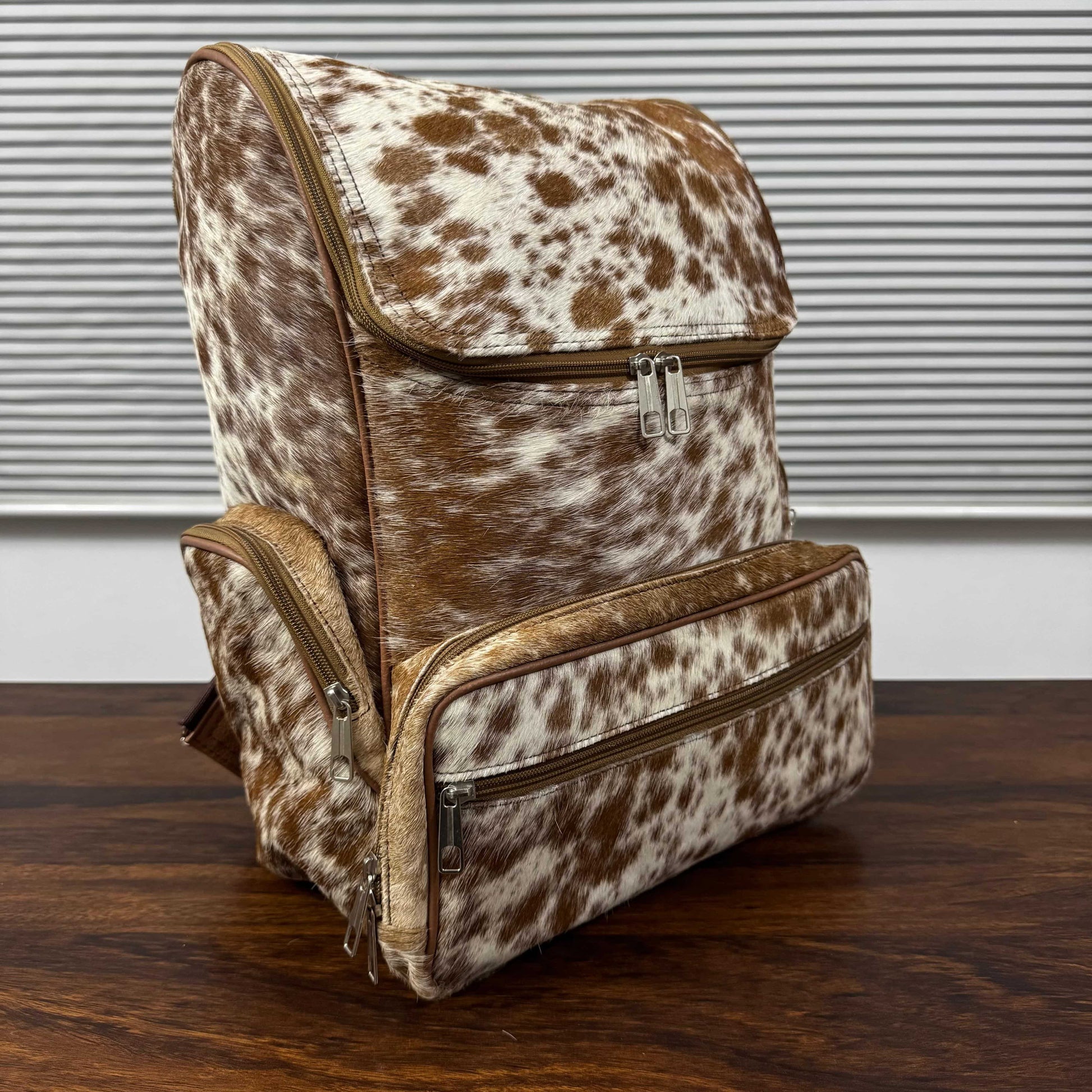 A cowhide-patterned backpack with brown and white spots, featuring tan leather trim and silver zippers, displayed on a dark wooden table against a background of gray horizontal blinds.