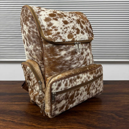A cowhide-patterned backpack with brown and white spots, featuring tan leather trim and silver zippers, displayed on a dark wooden table against a background of gray horizontal blinds.