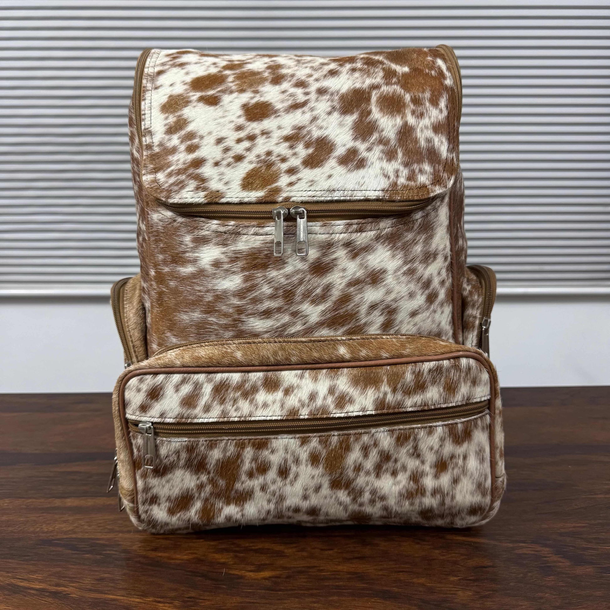 A brown and white cowhide-patterned backpack with matching faux-leather trim, shown standing on a dark wooden table in front of a window with horizontal blinds.