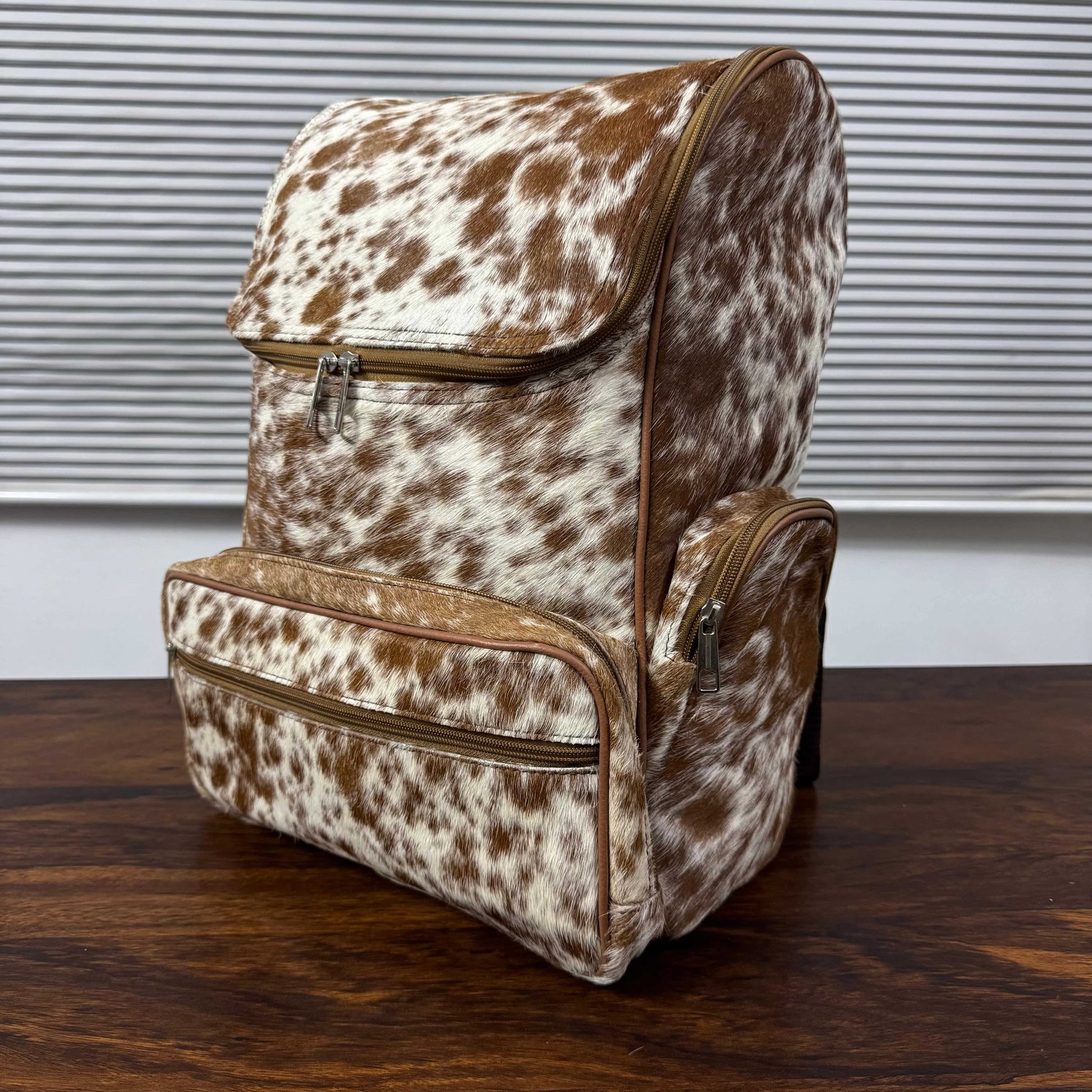 A backpack and matching diaper bag set in brown and white cowhide print, displayed on a dark wooden table in front of a window with horizontal blinds.
