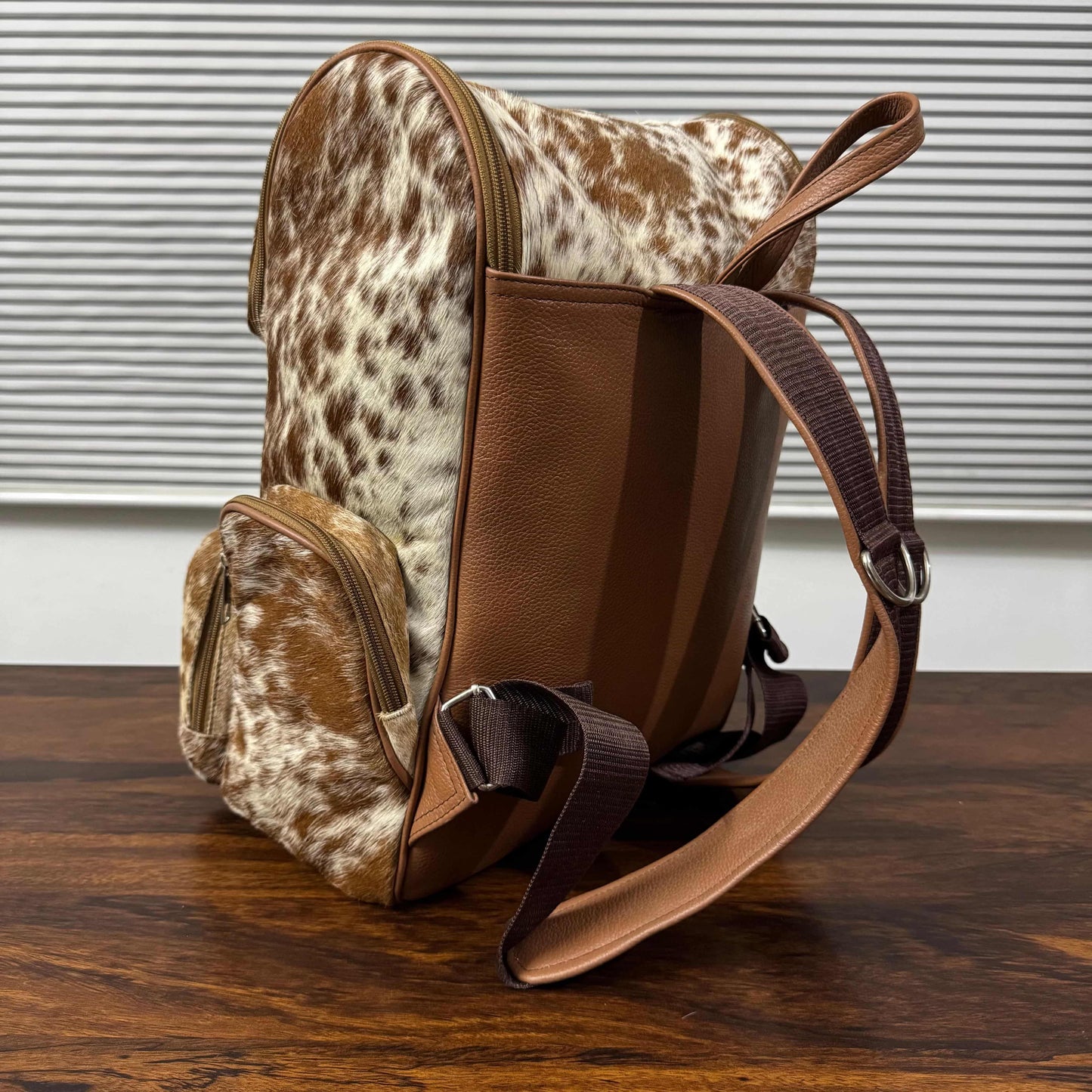 A brown and white cowhide-patterned backpack with smooth brown leather trim and a wide adjustable shoulder strap, shown standing on a wooden table in front of a window with horizontal blinds.