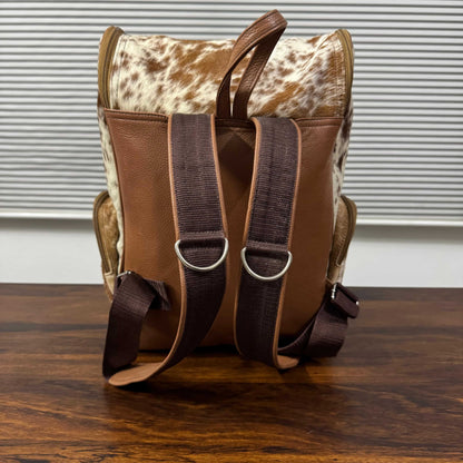 A brown leather and cowhide backpack shown from the back, resting on a dark wooden table in front of a window with blinds.