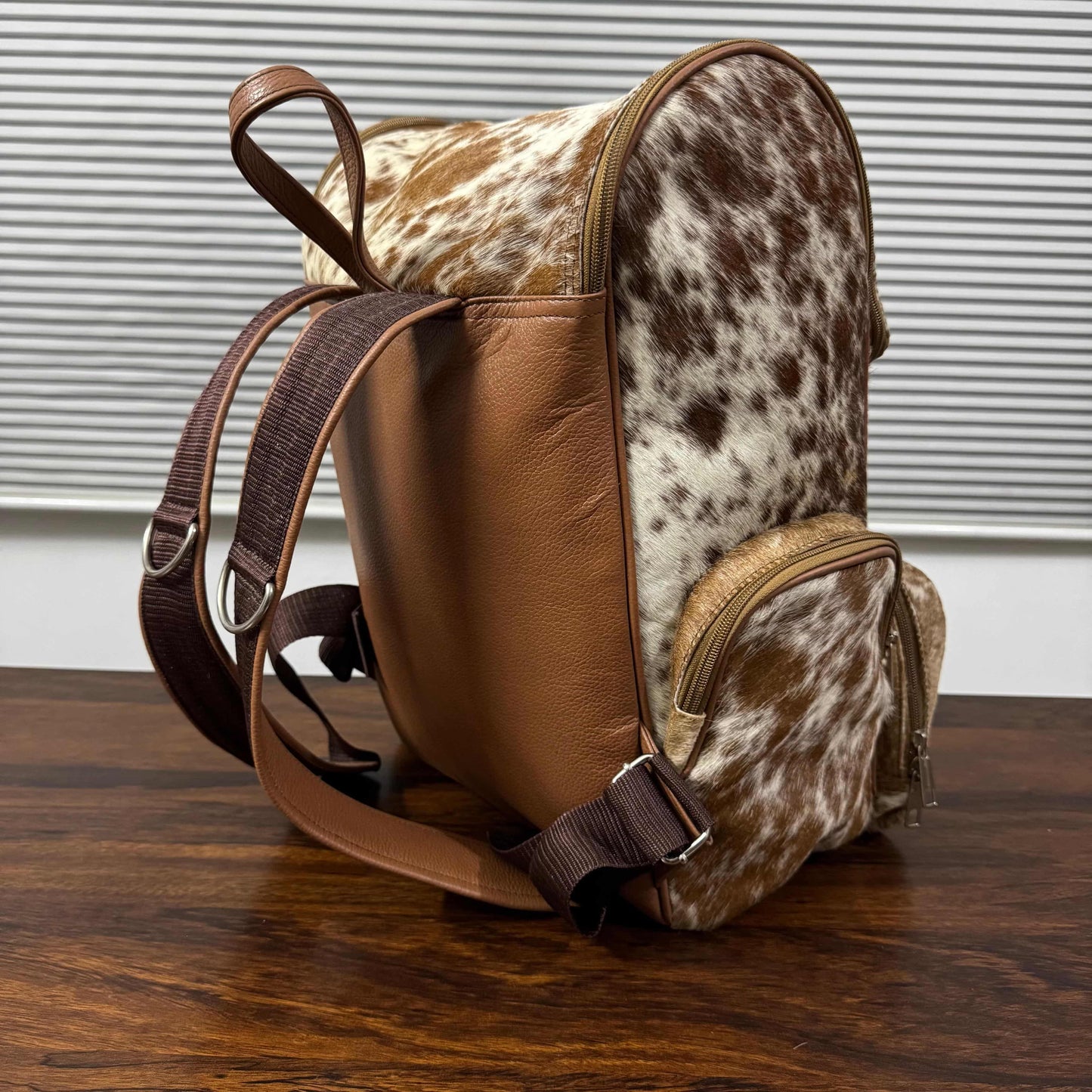 A brown and cowhide-patterned backpack resting on a dark wooden table in front of a window with horizontal blinds.