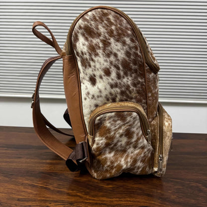 A small backpack with a brown and white cowhide pattern body, tan leather trim, and a brown shoulder strap, standing on a dark wooden surface in front of a window with horizontal blinds.