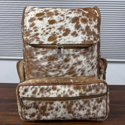 A brown and white cowhide-patterned backpack with a front flap pocket and a matching zippered front organizer pouch, displayed on a wooden surface against a gray slatted window background.