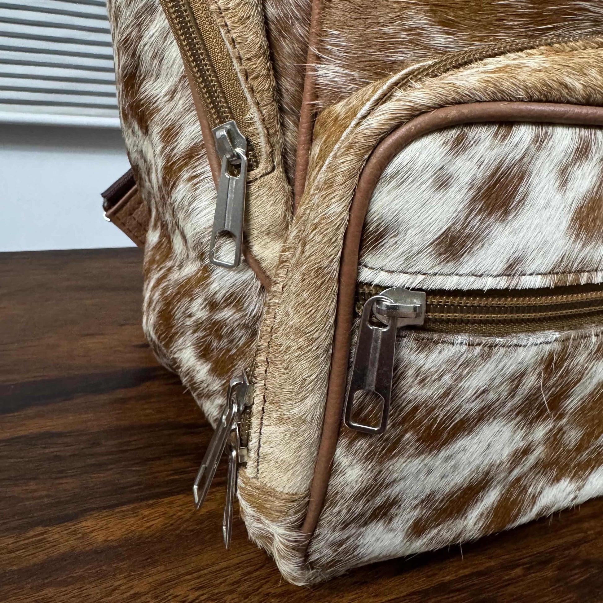 Close-up of a brown and white cowhide backpack or satchel with brown leather trim and silver-tone metal zippers, resting on a dark wood tabletop.