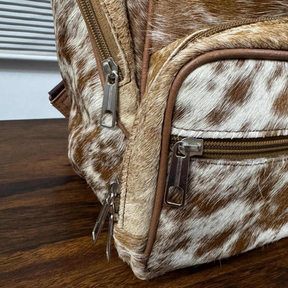 Close-up of a brown and white cowhide backpack or satchel with brown leather trim and silver-tone metal zippers, resting on a dark wood tabletop.