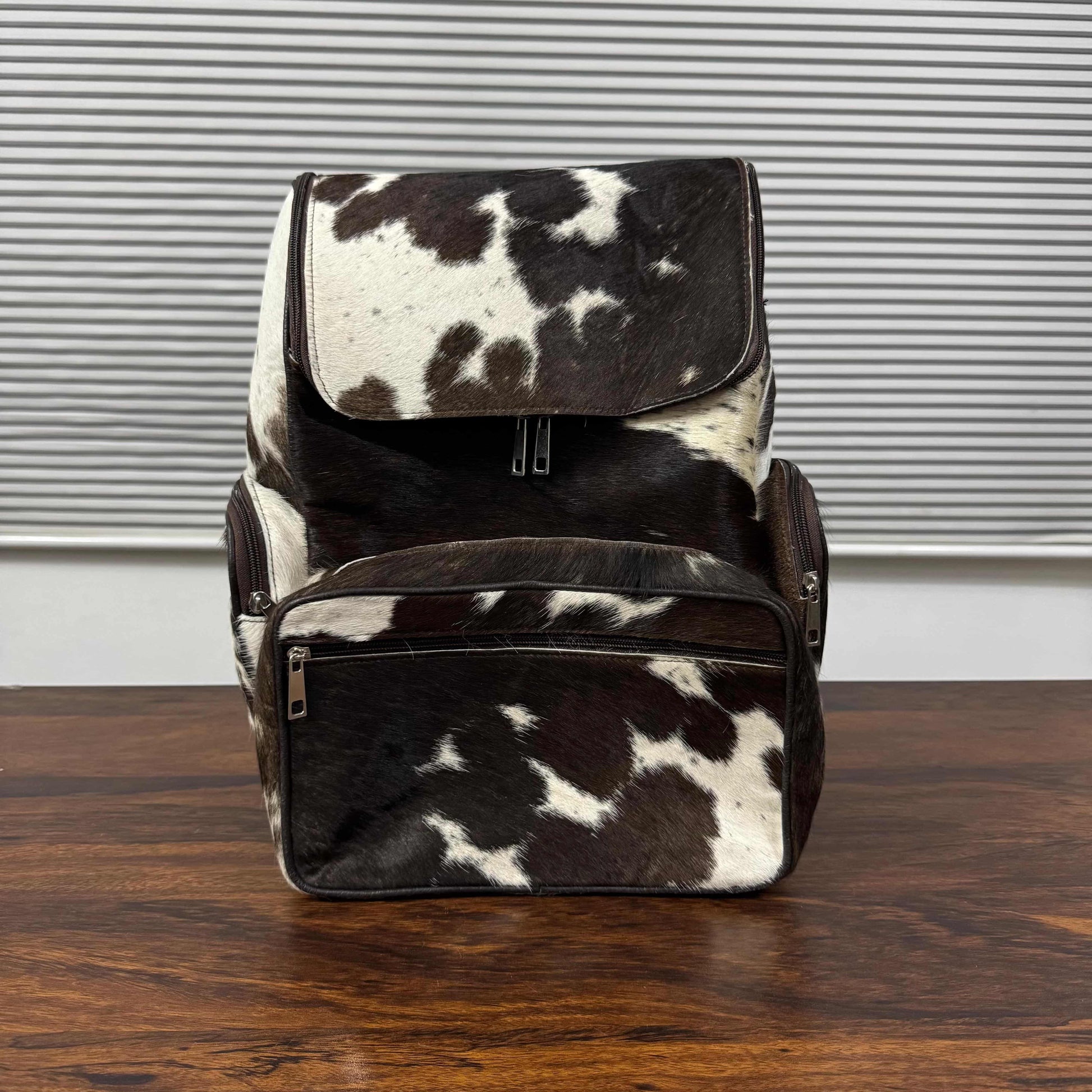 A cowhide backpack with a black and white spotted pattern, shown standing on a wooden table in front of a window with horizontal blinds.