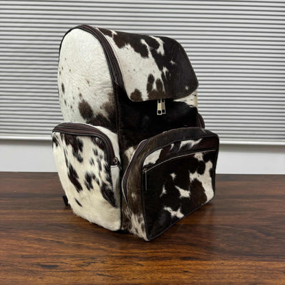 A cowhide backpack and matching zippered pouch with a black and white spotted pattern, displayed on a wooden table in front of a window with horizontal blinds.
