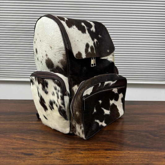A cowhide backpack and matching zippered pouch with a black and white spotted pattern, displayed on a wooden table in front of a window with horizontal blinds.