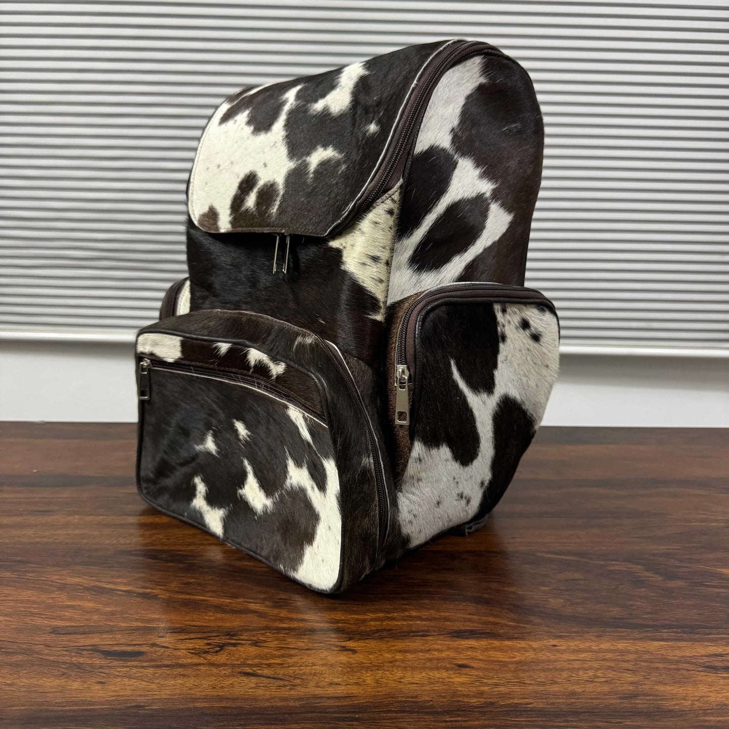 A cowhide backpack with a black and white spotted pattern, featuring a front zippered pocket and a side pocket, resting on a wooden table in front of a window with horizontal blinds.