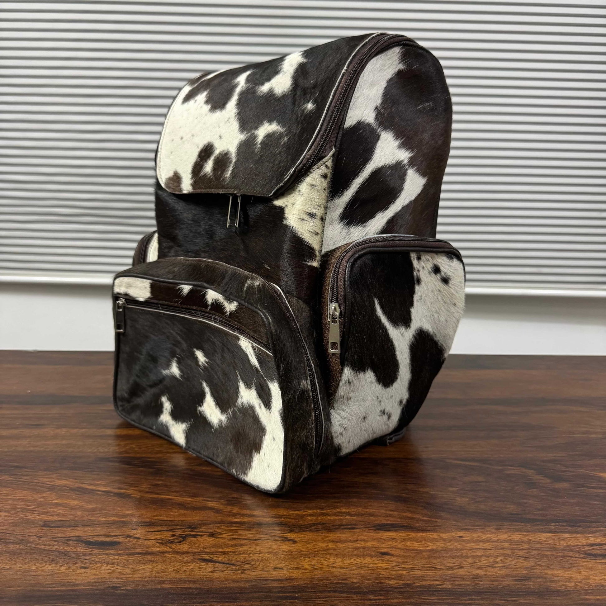 A cowhide backpack with a black and white spotted pattern, featuring a front zippered pocket and a side pocket, resting on a wooden table in front of a window with horizontal blinds.