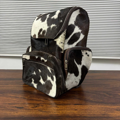 A cowhide backpack with a black and white spotted pattern, featuring a front zippered pocket and a side pocket, resting on a wooden table in front of a window with horizontal blinds.
