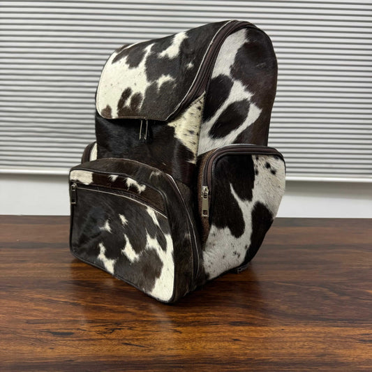 A cowhide backpack with a black and white spotted pattern, featuring a front zippered pocket and a side pocket, resting on a wooden table in front of a window with horizontal blinds.