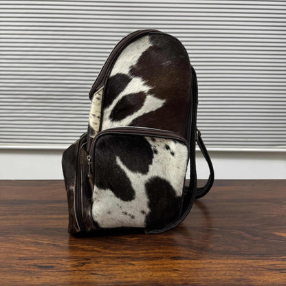 A cowhide backpack with a black and white spotted pattern, standing on a dark wooden table in front of a window with horizontal blinds.