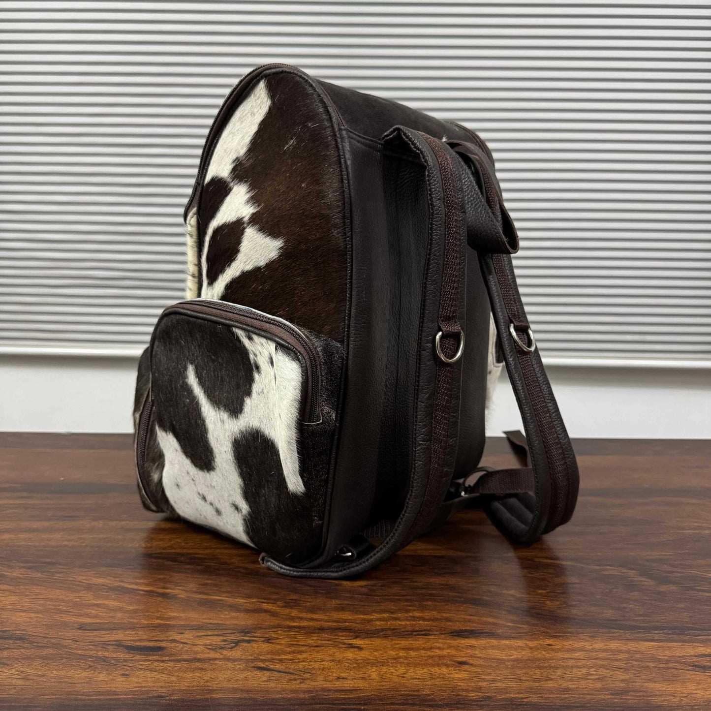 A dark brown leather and cowhide backpack with a black and white cow print pattern, resting on a wooden table in front of a window with horizontal blinds.