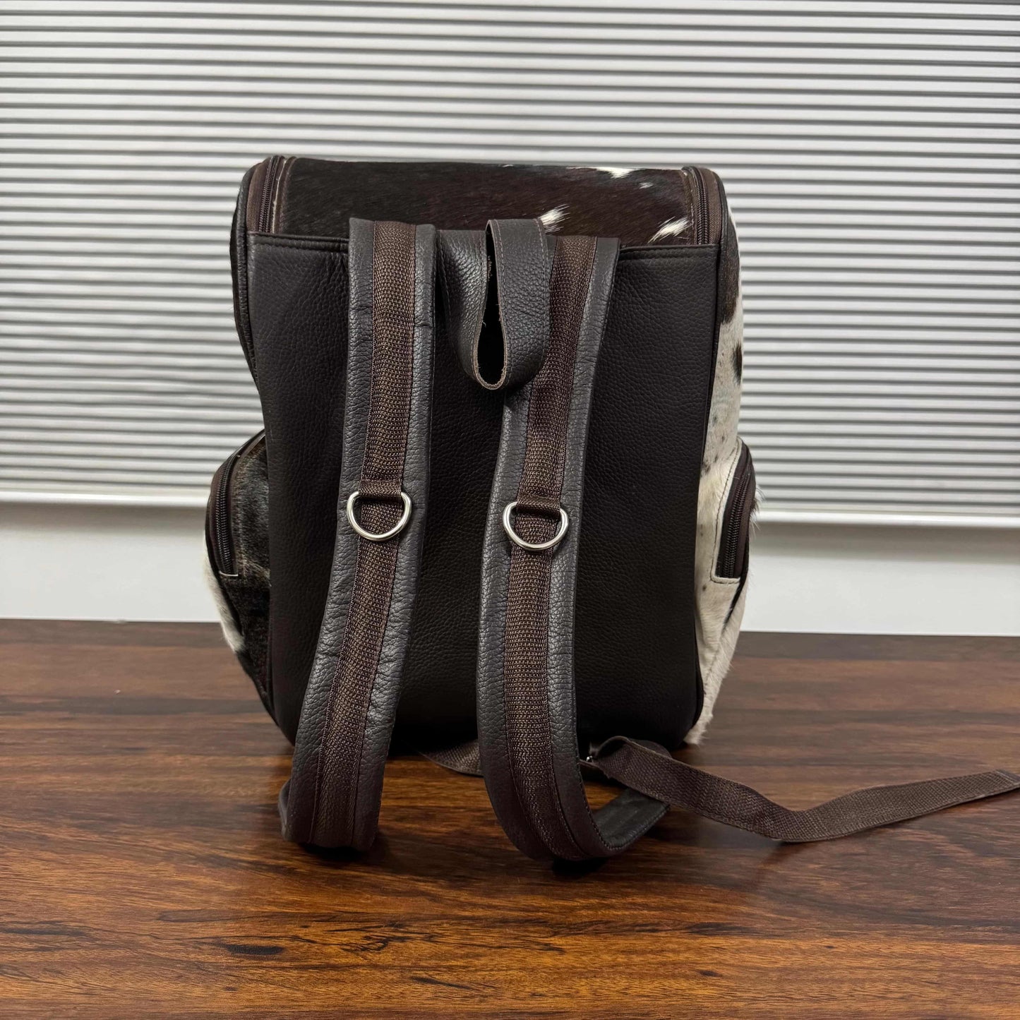 A dark brown leather backpack with cowhide-patterned side panels and matching brown leather shoulder straps, standing on a wooden table in front of a window with horizontal blinds.