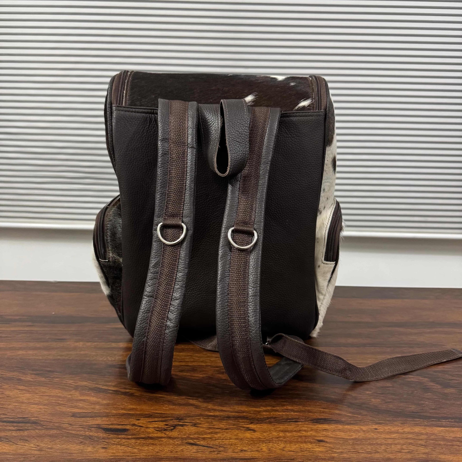 A dark brown leather backpack with cowhide-patterned side panels and matching brown leather shoulder straps, standing on a wooden table in front of a window with horizontal blinds.