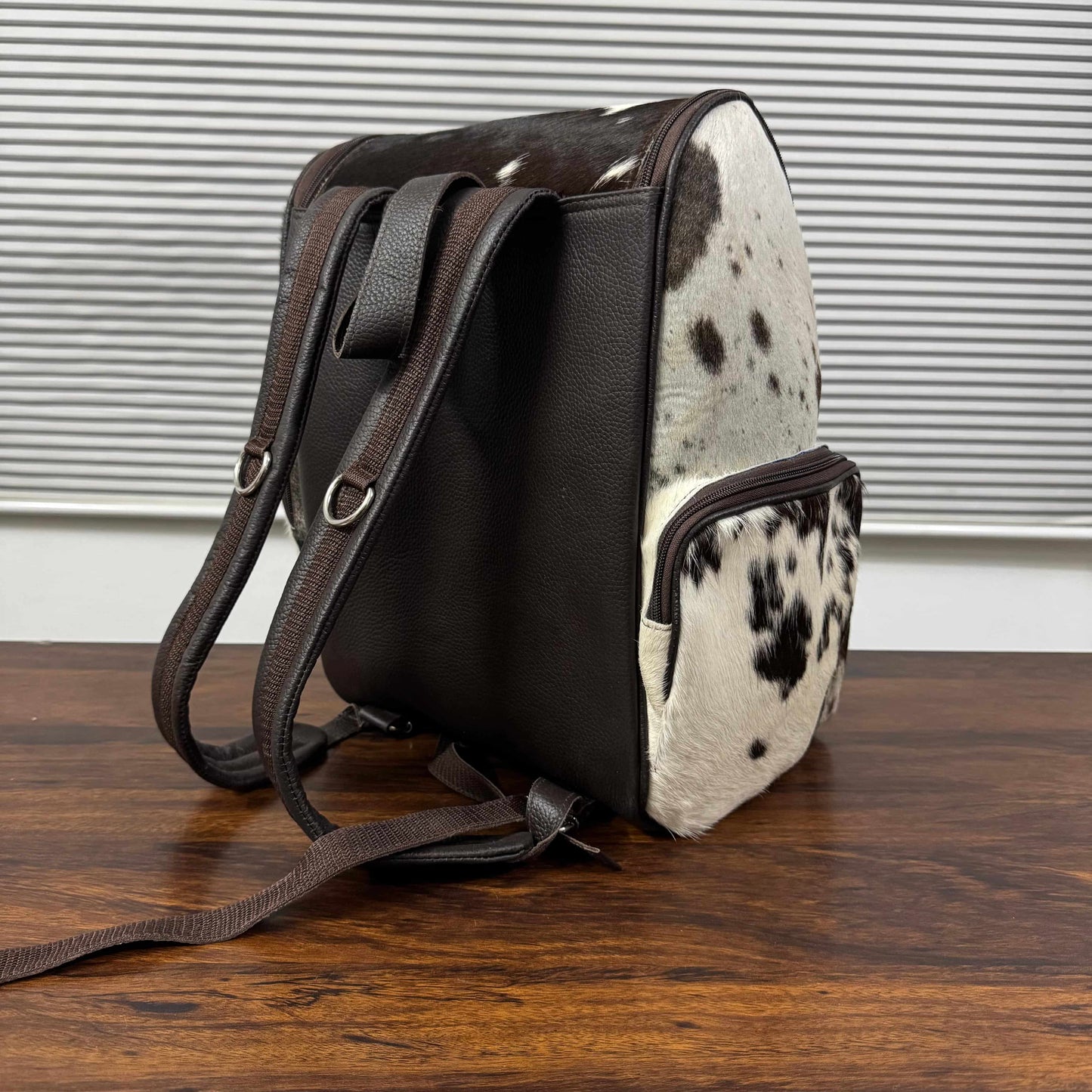 A small shoulder bag with dark brown faux-leather panels and a black-and-white cowhide patterned front and side, shown standing on a wooden surface against a window-blind background.