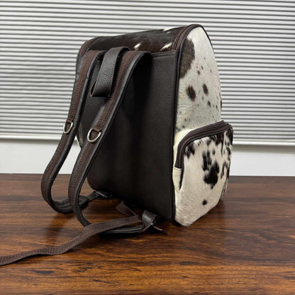 A small shoulder bag with dark brown faux-leather panels and a black-and-white cowhide patterned front and side, shown standing on a wooden surface against a window-blind background.
