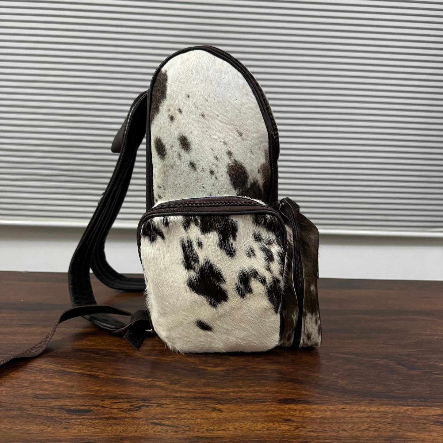 A small shoulder bag with a black and white cowhide pattern and dark brown leather trim, standing on a wooden table in front of a gray slatted wall.