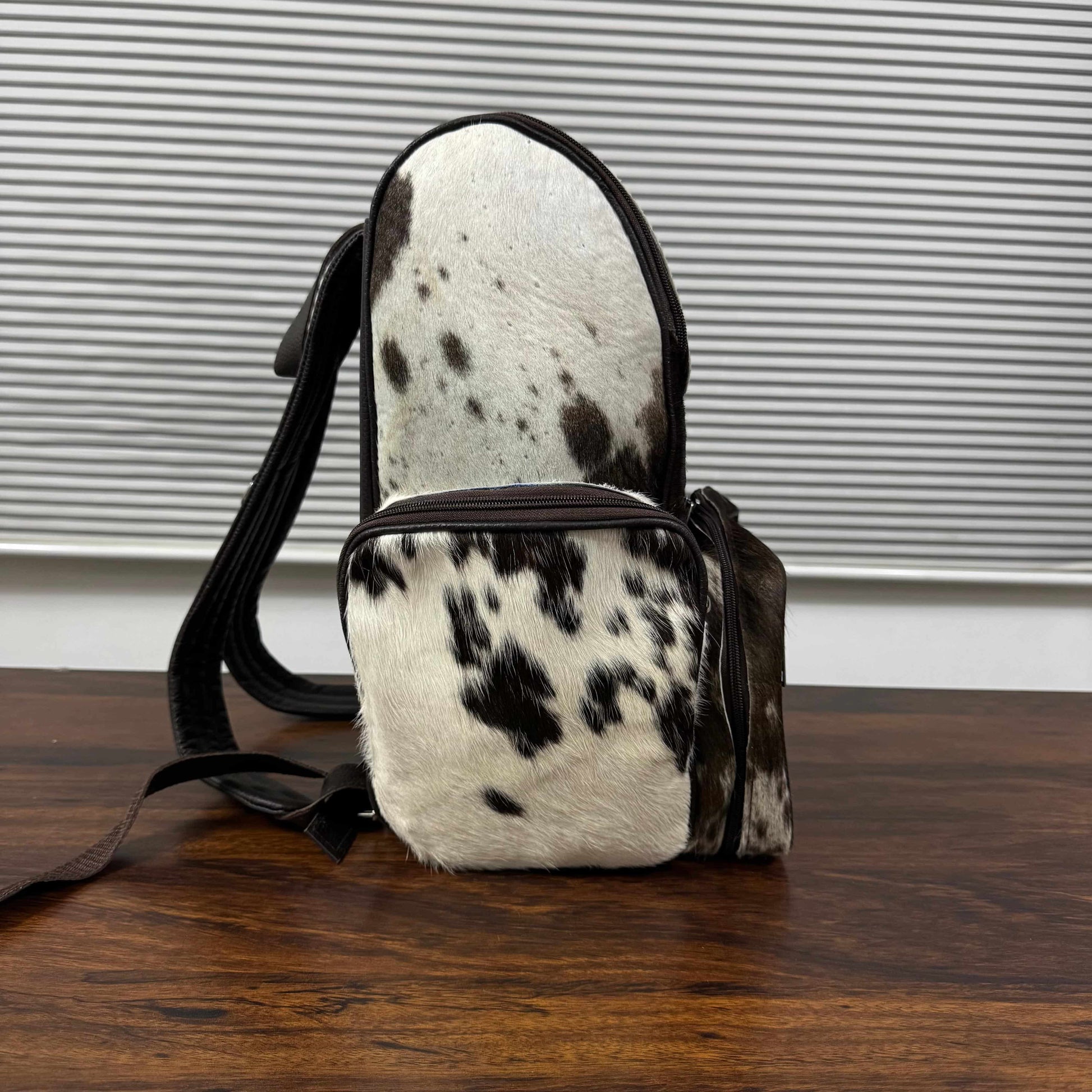 A small shoulder bag with a black and white cowhide pattern and dark brown leather trim, standing on a wooden table in front of a gray slatted wall.