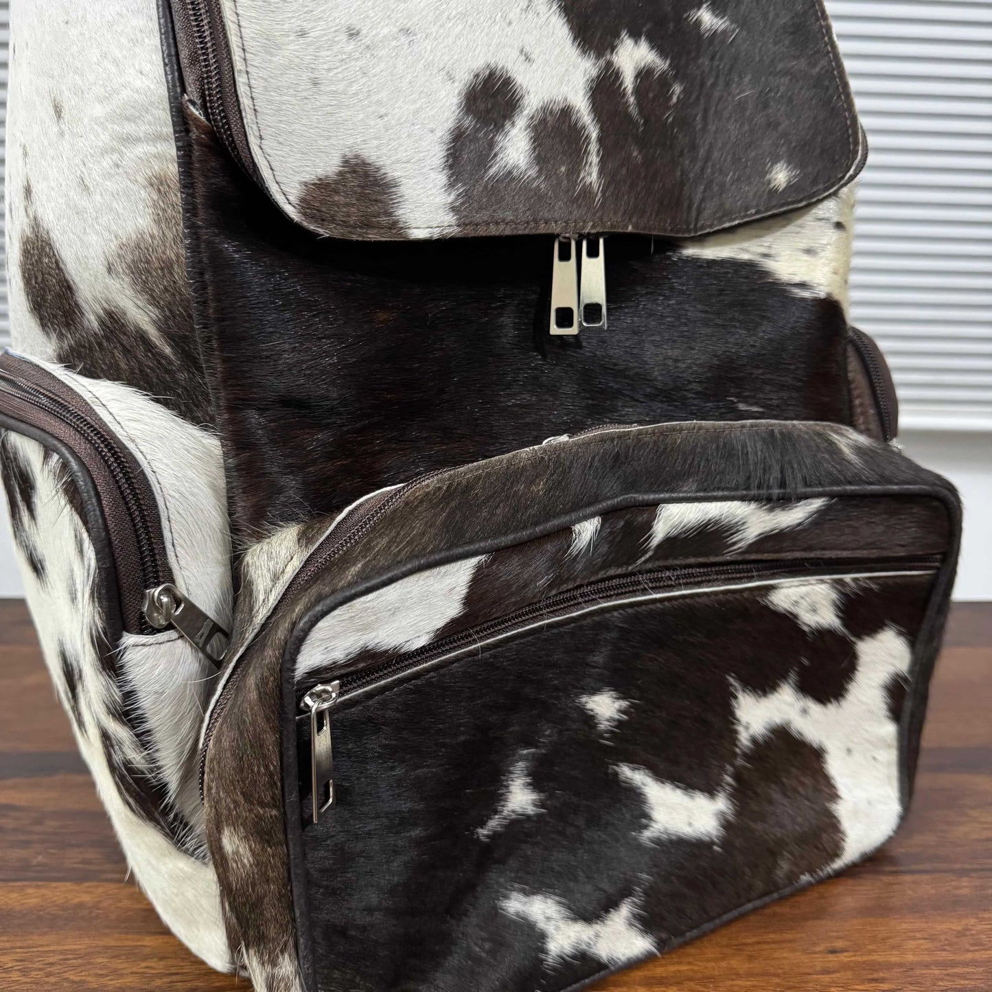A black and white cowhide backpack with dark brown trim, shown on a wooden surface with a window blinds background.
