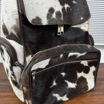 A black and white cowhide backpack with dark brown trim, shown on a wooden surface with a window blinds background.