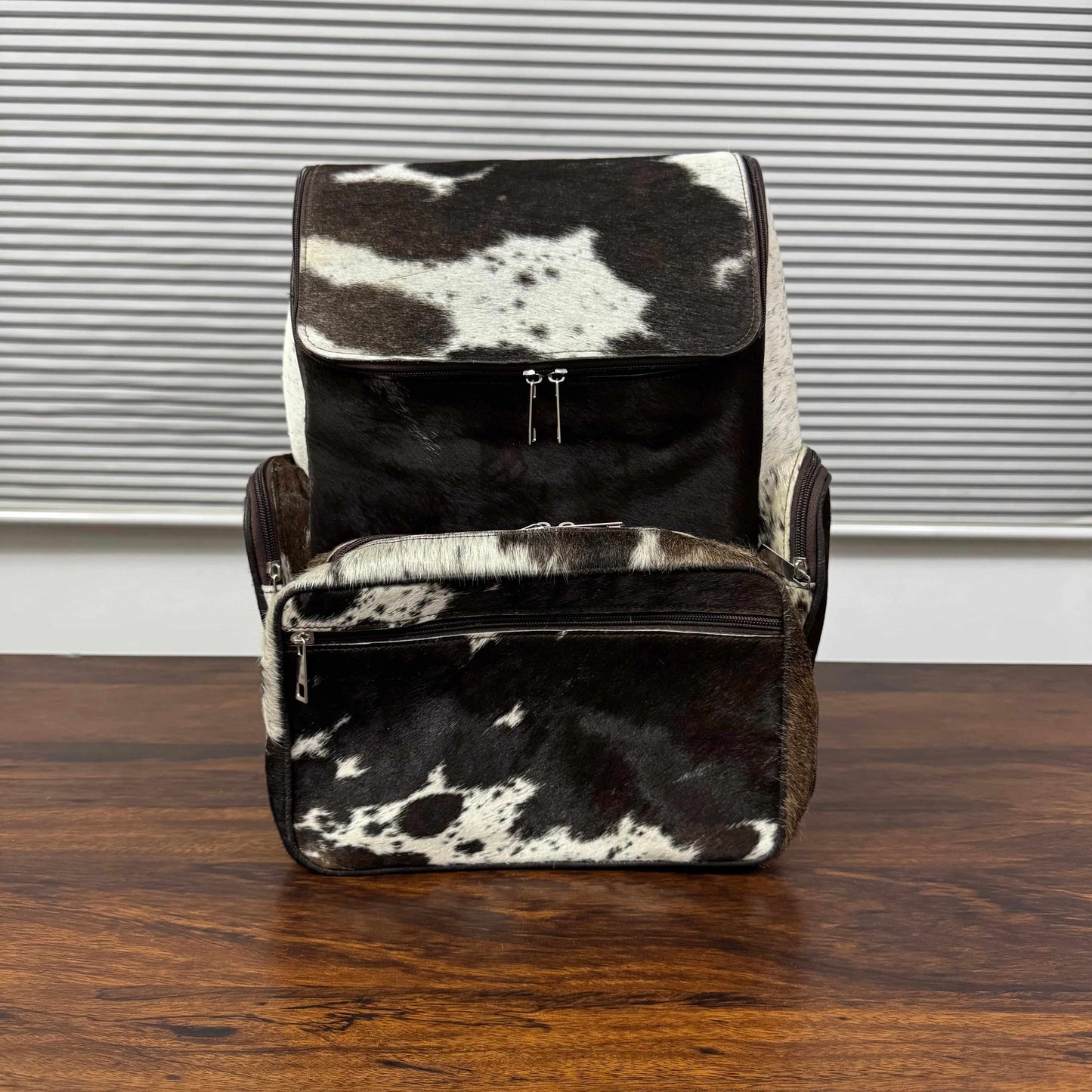 A black and white cowhide backpack and matching zippered pouch set on a dark wooden table in front of a window with horizontal blinds.