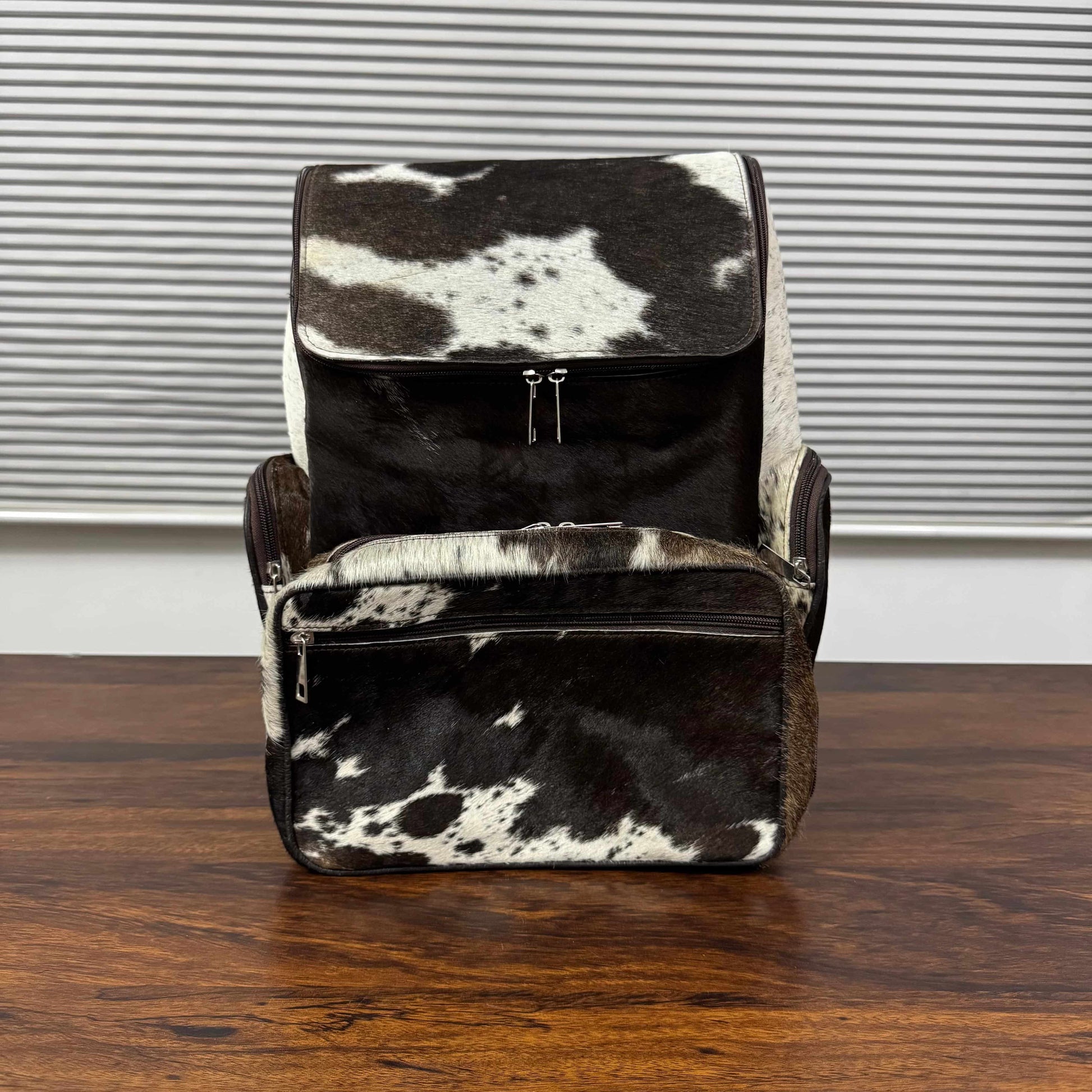 A black and white cowhide backpack and matching zippered pouch set on a dark wooden table in front of a window with horizontal blinds.