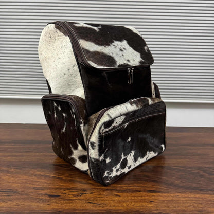 A backpack and matching zip pouch set in black and white cowhide, displayed on a dark wooden table in front of a window with horizontal blinds.