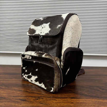 A cowhide backpack with a black and white spotted pattern, featuring a front zippered pouch and a main compartment, displayed on a wooden table in front of a window with horizontal blinds.