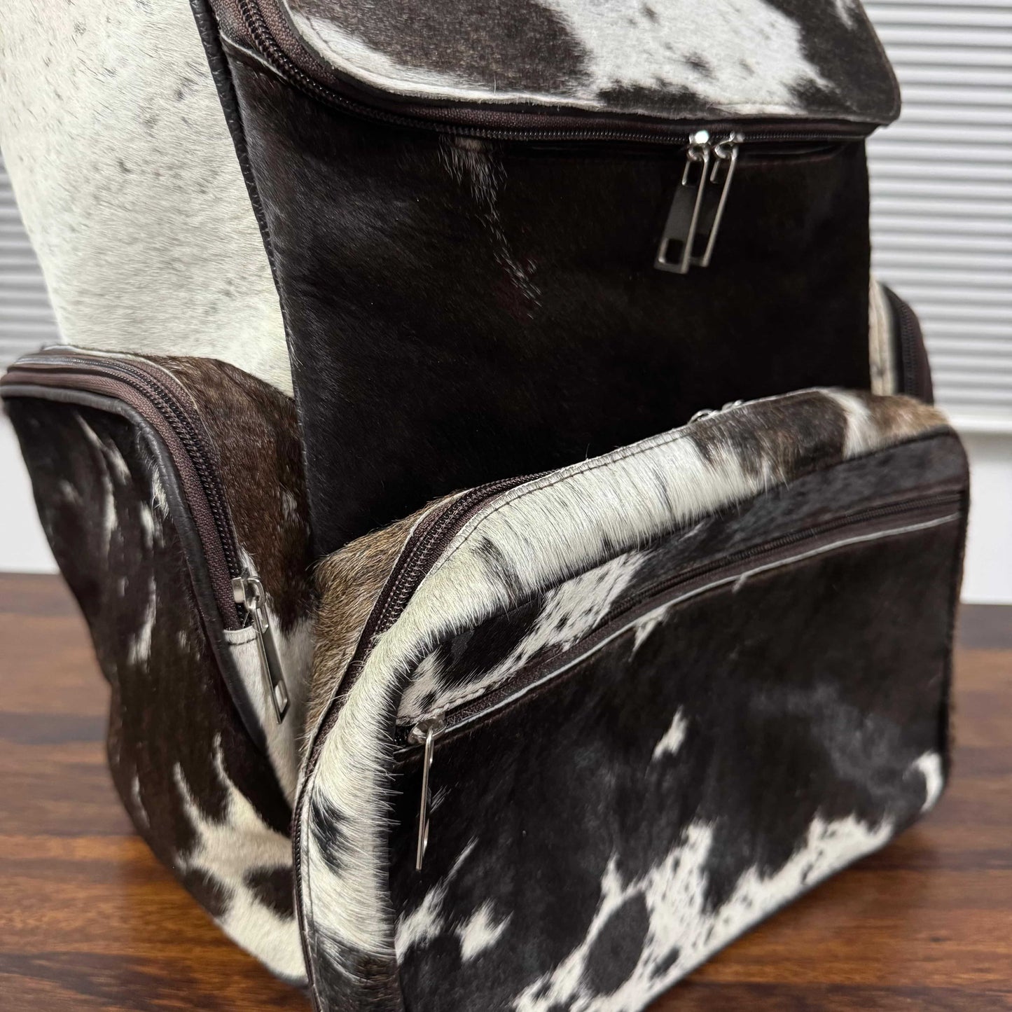A structured backpack and matching zippered pouch in a black and white cowhide pattern, displayed on a wooden surface with a window blinds background.