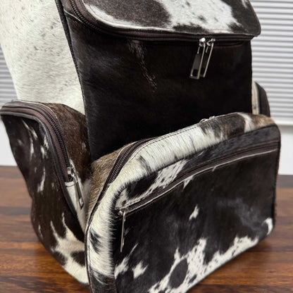 A structured backpack and matching zippered pouch in a black and white cowhide pattern, displayed on a wooden surface with a window blinds background.