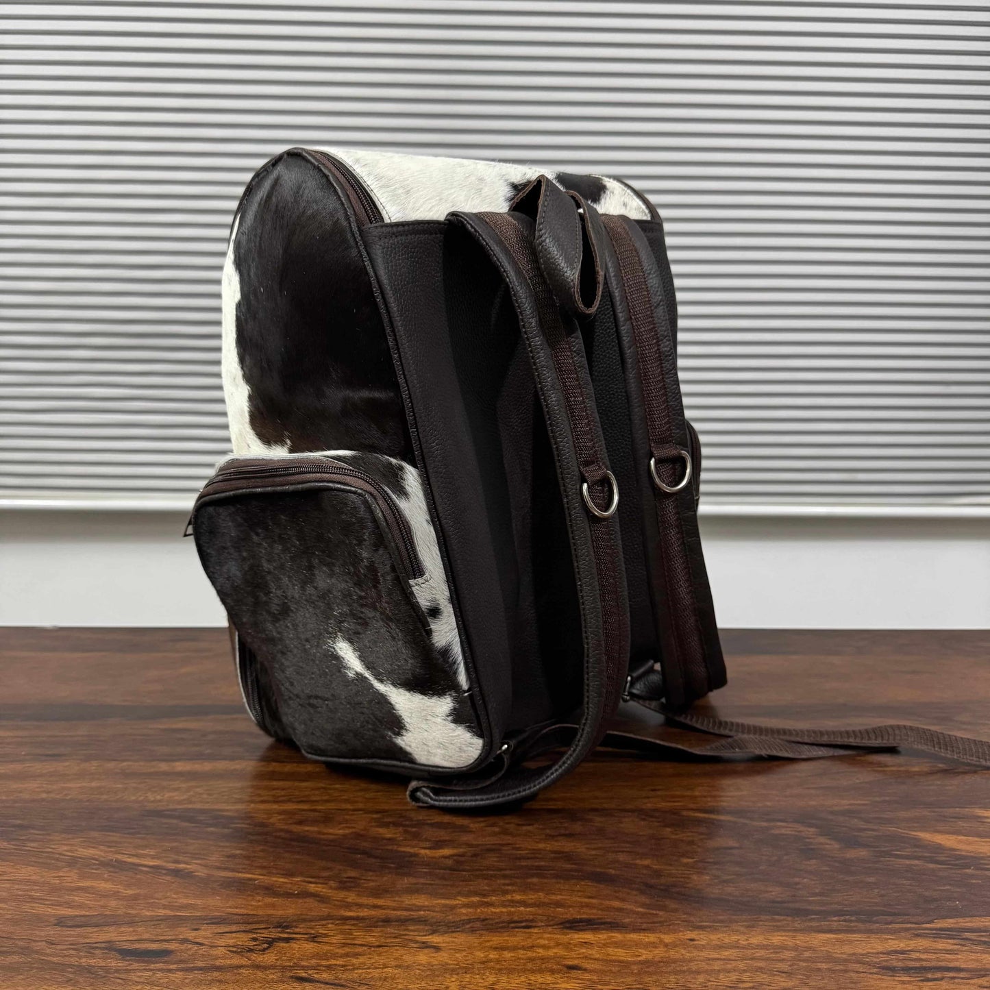 A cowhide and dark brown leather backpack standing upright on a wooden table in front of a window with horizontal blinds.