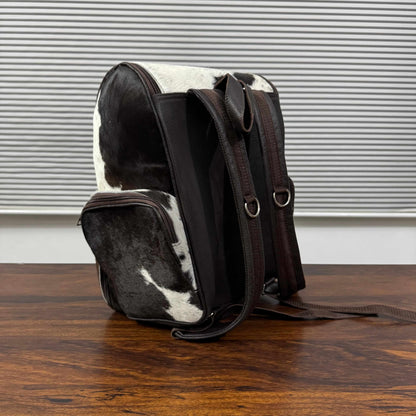 A cowhide and dark brown leather backpack standing upright on a wooden table in front of a window with horizontal blinds.