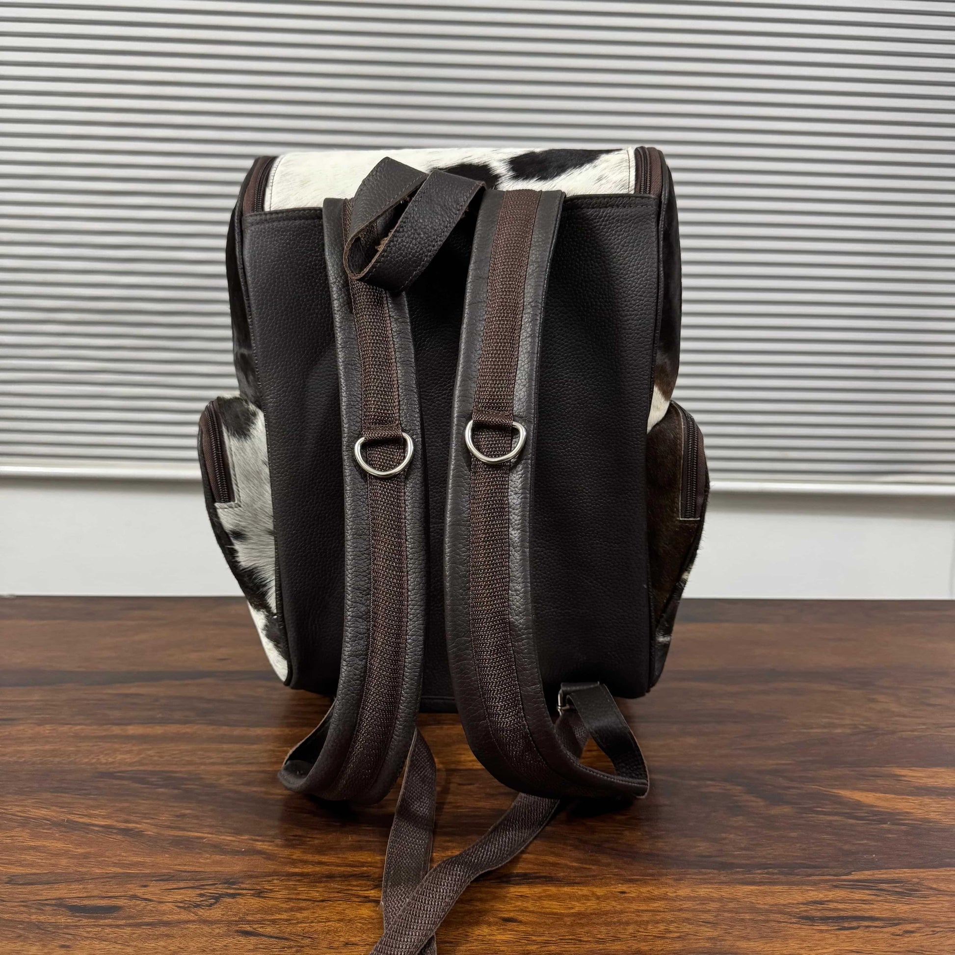 A black and brown leather backpack with cowhide accents, shown from the back on a wooden table in front of a gray slatted window.