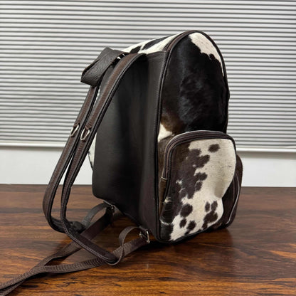 A cowhide and leather backpack resting on a dark wooden table in front of a gray slatted wall. The backpack features a black and white cowhide body, dark brown leather straps and trim, and a front zippered pocket.