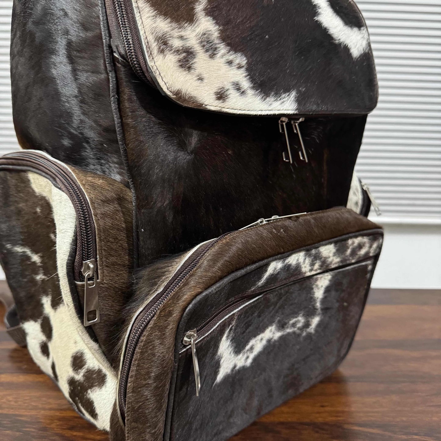 A structured backpack and matching diaper bag set made from dark brown and cream cowhide, displayed on a wooden tabletop with a window blind in the background.